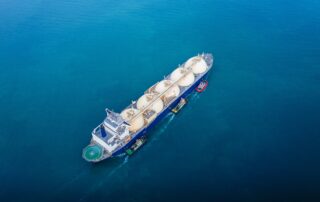 Aerial view of a large LNG (liquefied natural gas) tanker escorted by tugboats on calm blue water, highlighting marine transport of alternative fuels.