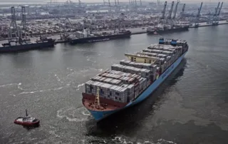 large Maersk Line container ship navigates a busy commercial port with the assistance of a tugboat. The vessel is loaded with stacked shipping containers, and multiple cranes and cargo terminals are visible in the background.