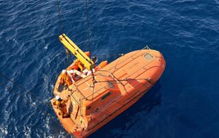 Seafarers in safety gear lowering an orange enclosed lifeboat into the sea during STCW survival craft training.