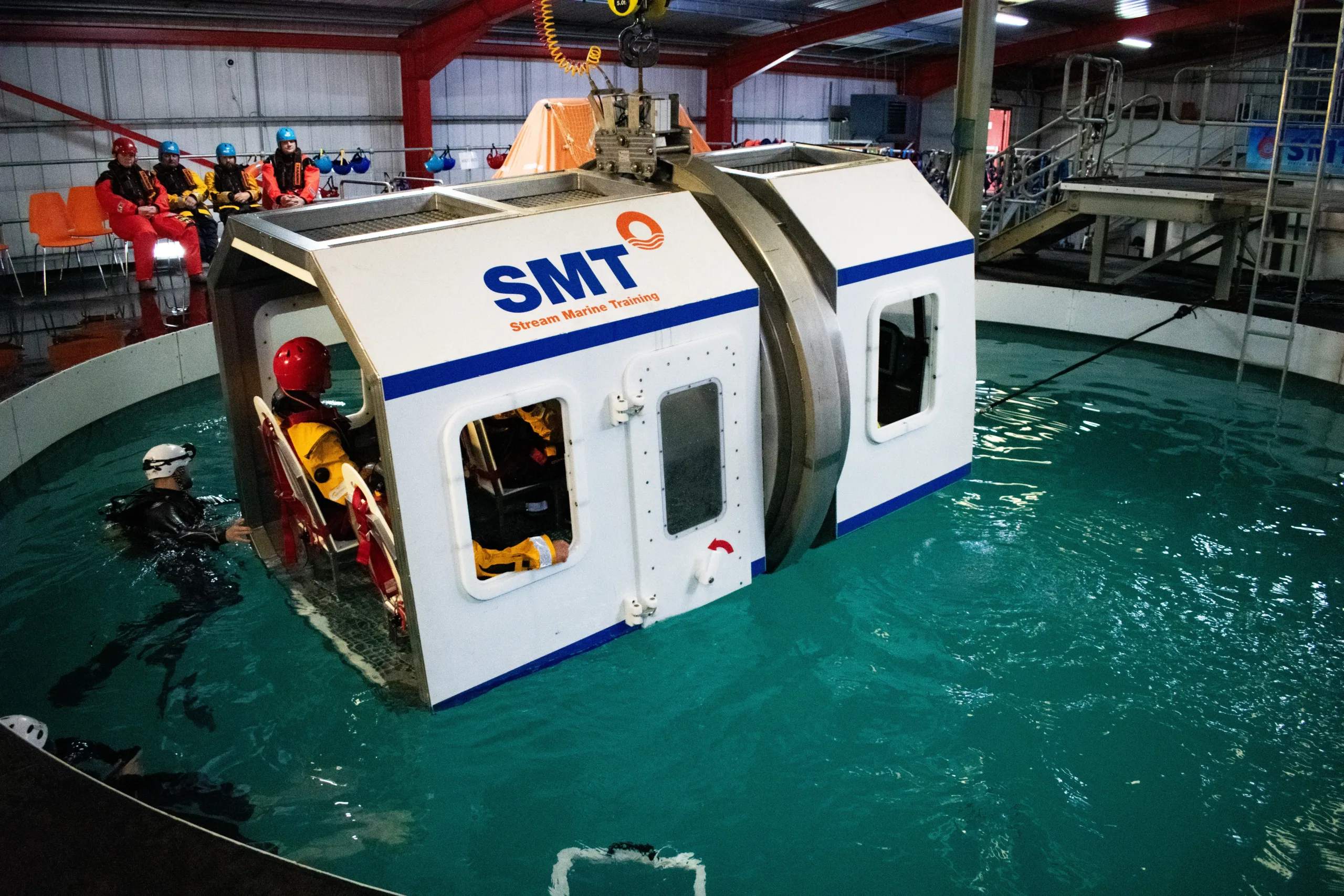 HUET simulator being lowered into pool while delegates in hi-vis and helmets are strapped inside.