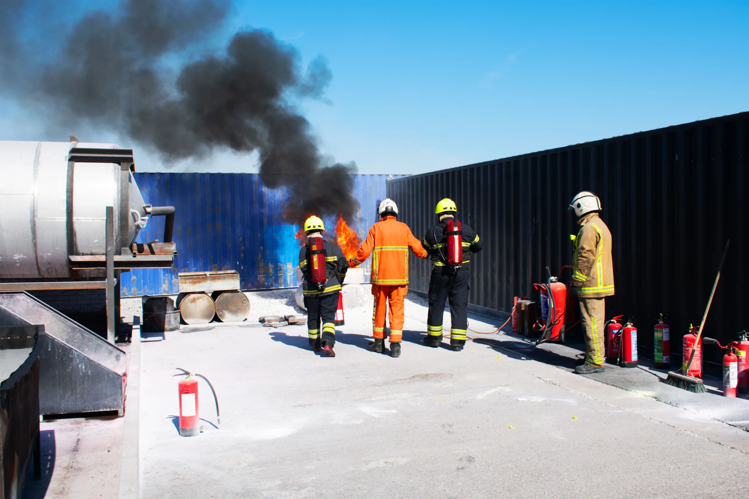 Stream marine fire instructor in orange fireproof clothing takes two delegates in similar attire and with ba-sets on with fire extinguishers towards and active fire billowing black smoke.