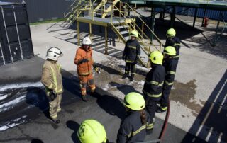 Offshore workers in firefighting gear gathered around an instructor during OPITO FOET refresher training at Stream Marine Training’s fire ground.