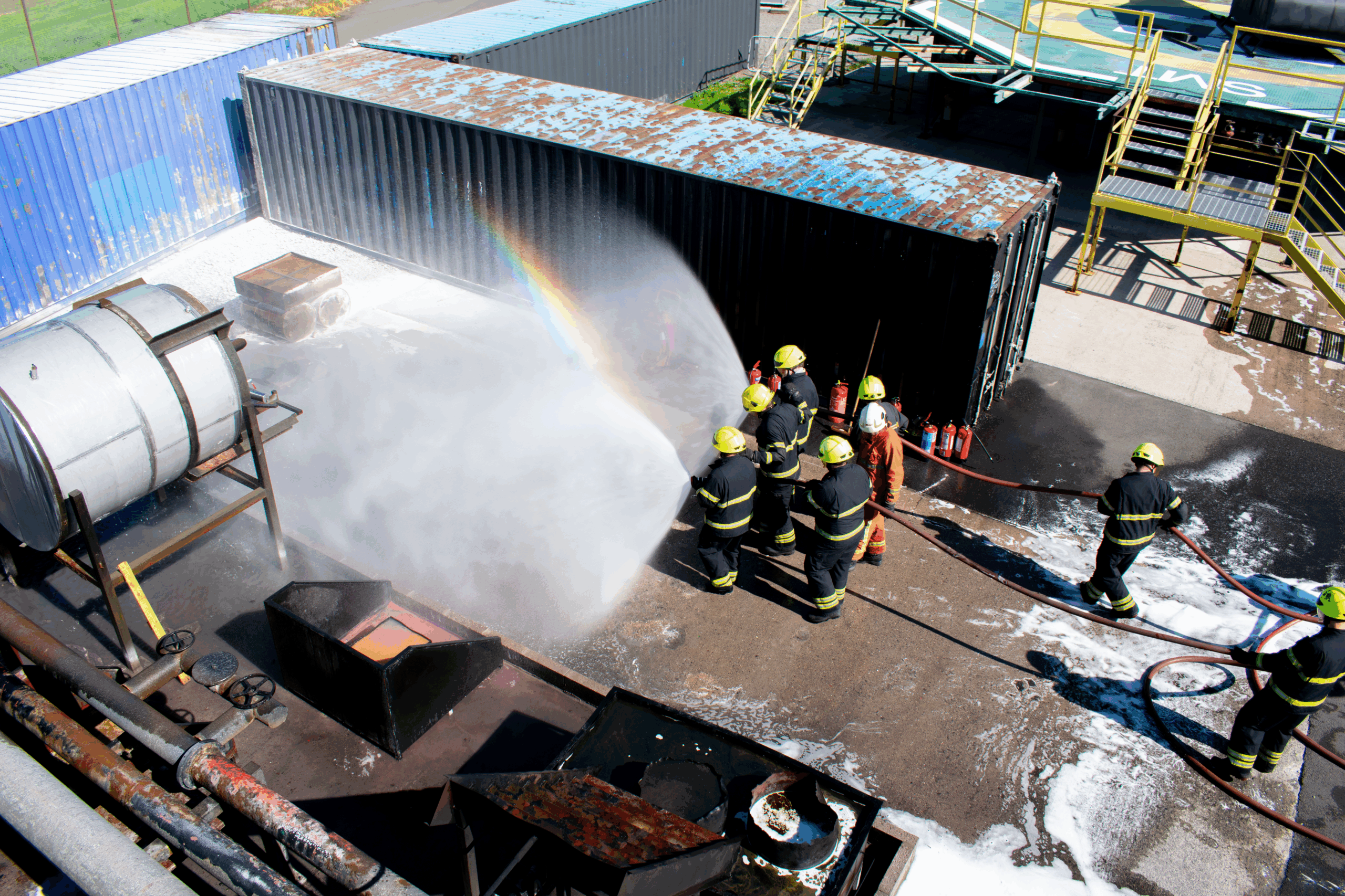 a group of firefighter delegates walk together with two hoses spray water at an active tanker fire